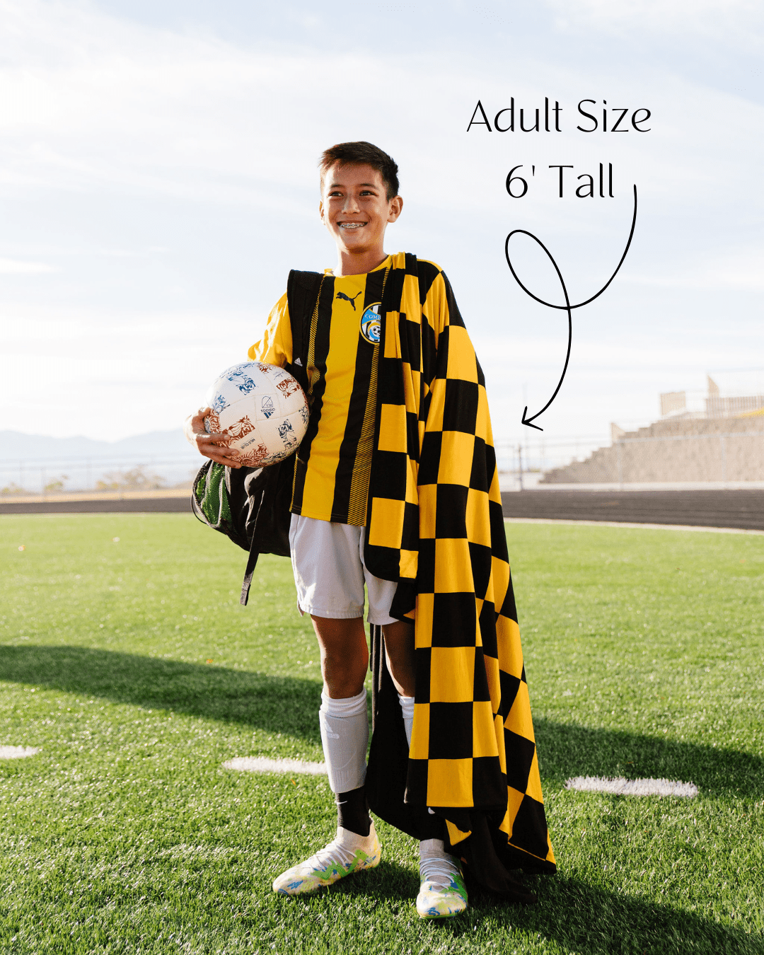 Soccer player wrapped in black and gold yellow checked gameday blanket showing that it is 6 feet tall, covering him from head to toe. 